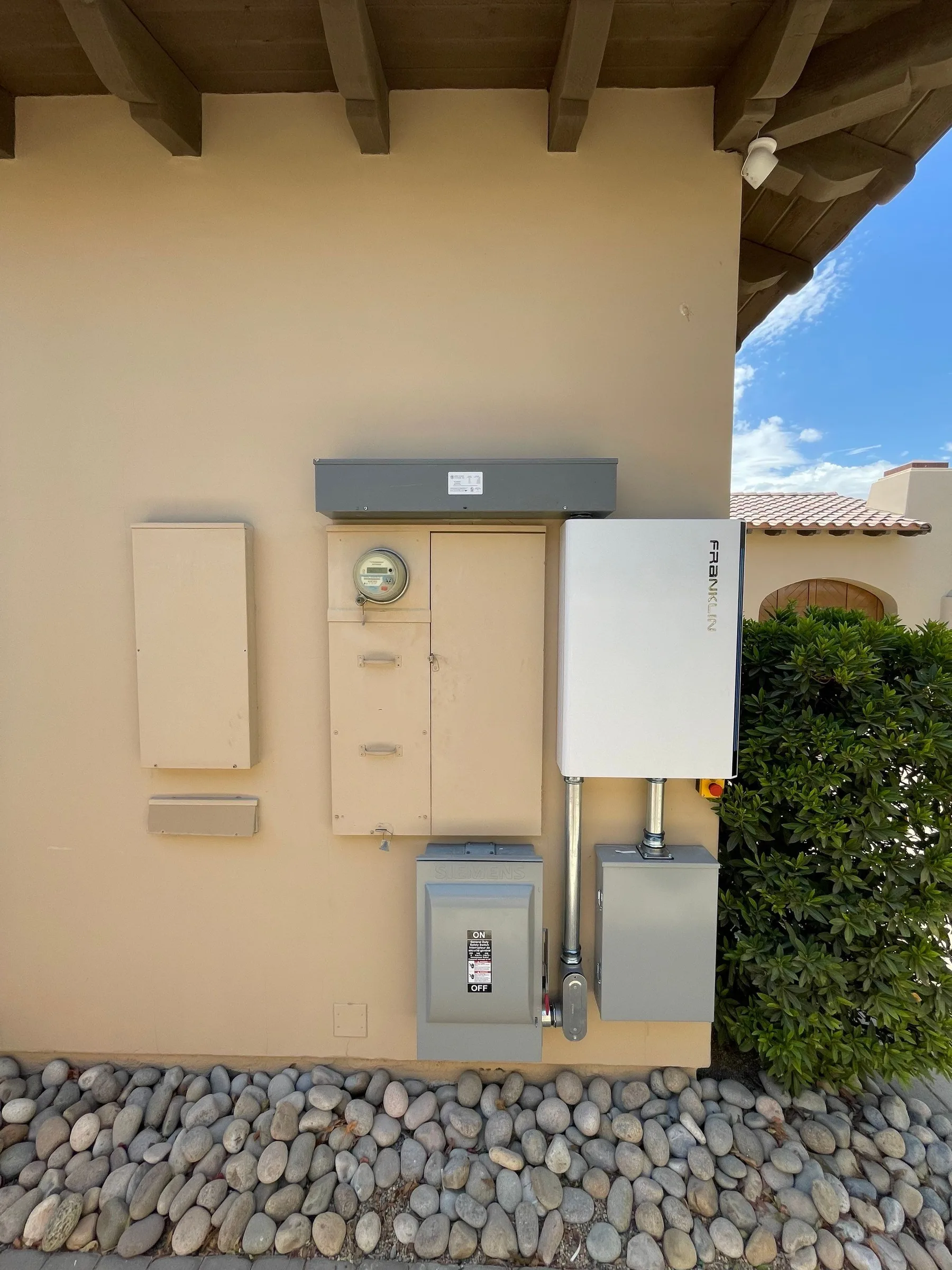 A Franklin control unit beside an outdoor meter panel and disconnects on a stucco wall under a tile roof.